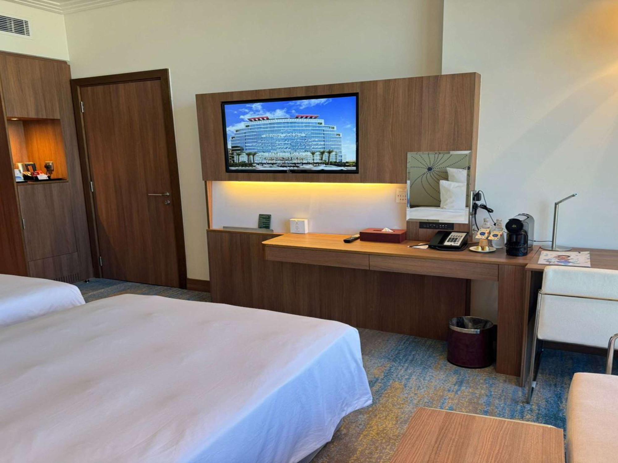 Novotel Jazan Exterior photo The photo shows a hotel room with two beds made up with white linens. There is a wooden wall unit that features a flat-screen television mounted on it, displaying a picture of a large building. Below the TV, there is a desk area with various items, i